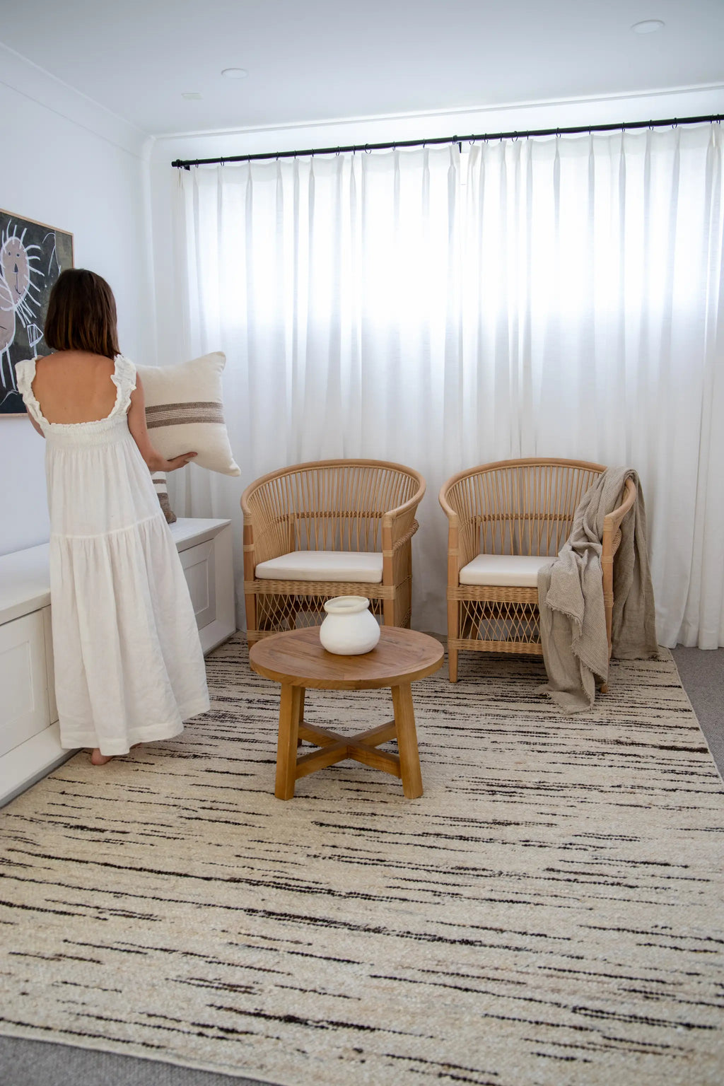 Woman in a white dress standing in a modern living room with wicker chairs and a wooden table on a wool rug from Rugs For Good. 