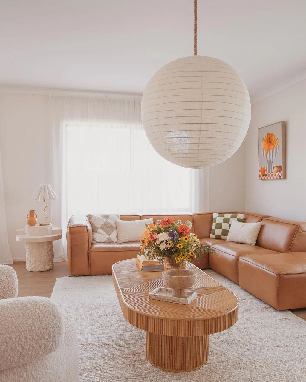 A bright and spacious living room with a hand knotted white wool rug on a brown leather sofa, complemented by a round wooden coffee table and a large spherical pendant light.
